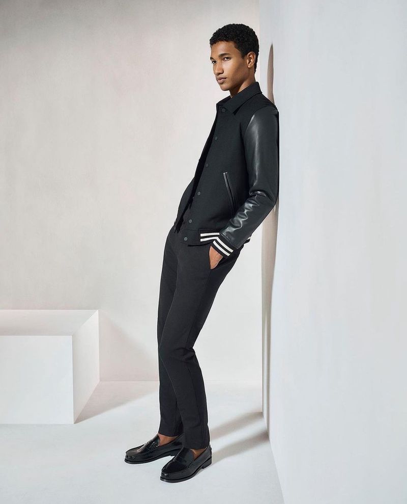 A young adult male model poses leaning against a wall in a black varsity jacket, trousers, and loafers in a studio setting.