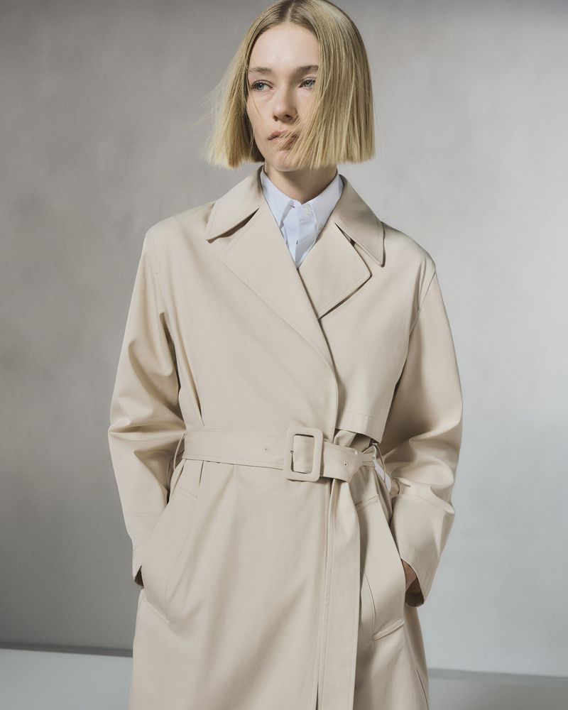 A fair-skinned woman with blonde bobbed hair is captured indoors wearing a tan trench coat and light blue shirt against a neutral backdrop.