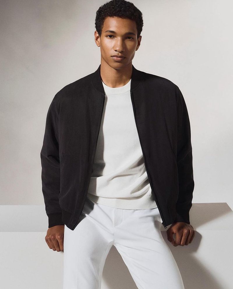 Adult male fashion model poses in a studio wearing a black jacket over a white shirt and white pants. He is looking at the camera.