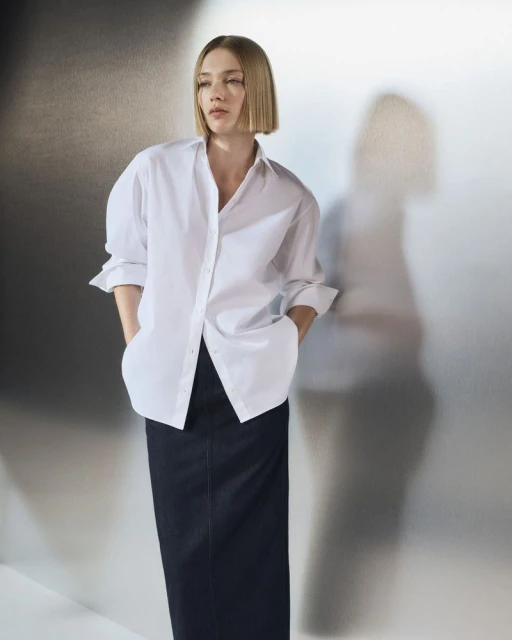 Adult female in a white shirt and black skirt poses in front of a textured backdrop in a fashion-style medium studio shot.
