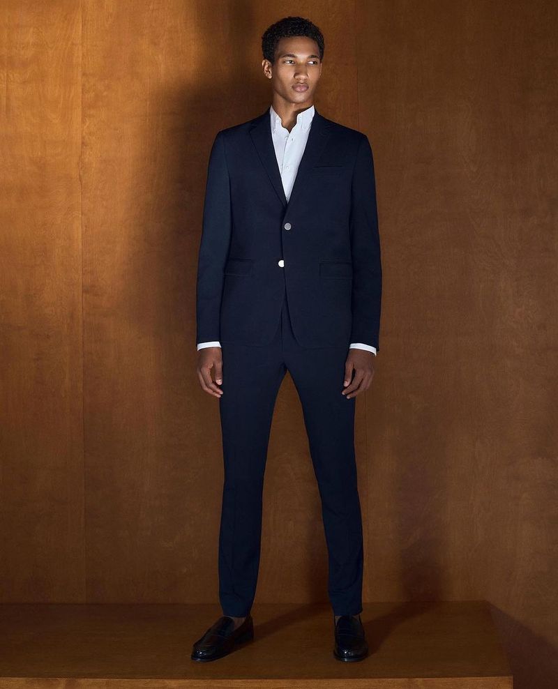 A young adult male model poses indoors against a bronze backdrop wearing a navy blue suit with a white shirt and black dress shoes.