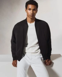 A black man wearing a black bomber jacket over a white top and white trousers against a neutral background.