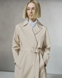Blonde model with a blunt bob haircut wearing a belted beige trench coat and collared shirt in a studio setting.