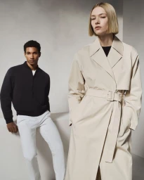 Two models posing against a neutral studio backdrop, featuring a woman in a cream trench coat and a man in a black jacket.