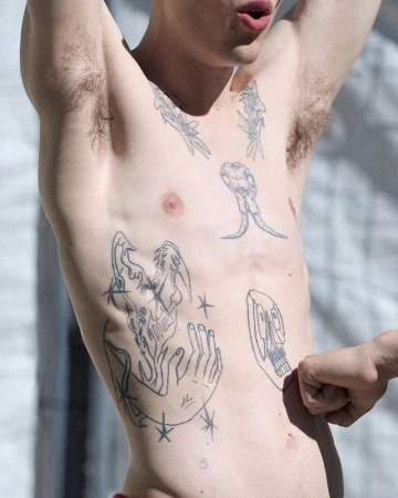 Close-up shot of a shirtless person with multiple tattoos on their torso and arms, raising their arms above their head.