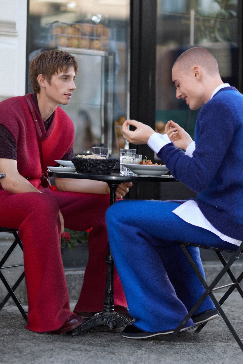 Two stylish people in red and blue outfits sit at an outdoor cafe table, enjoying a meal and engaging in conversation on a sunny day.