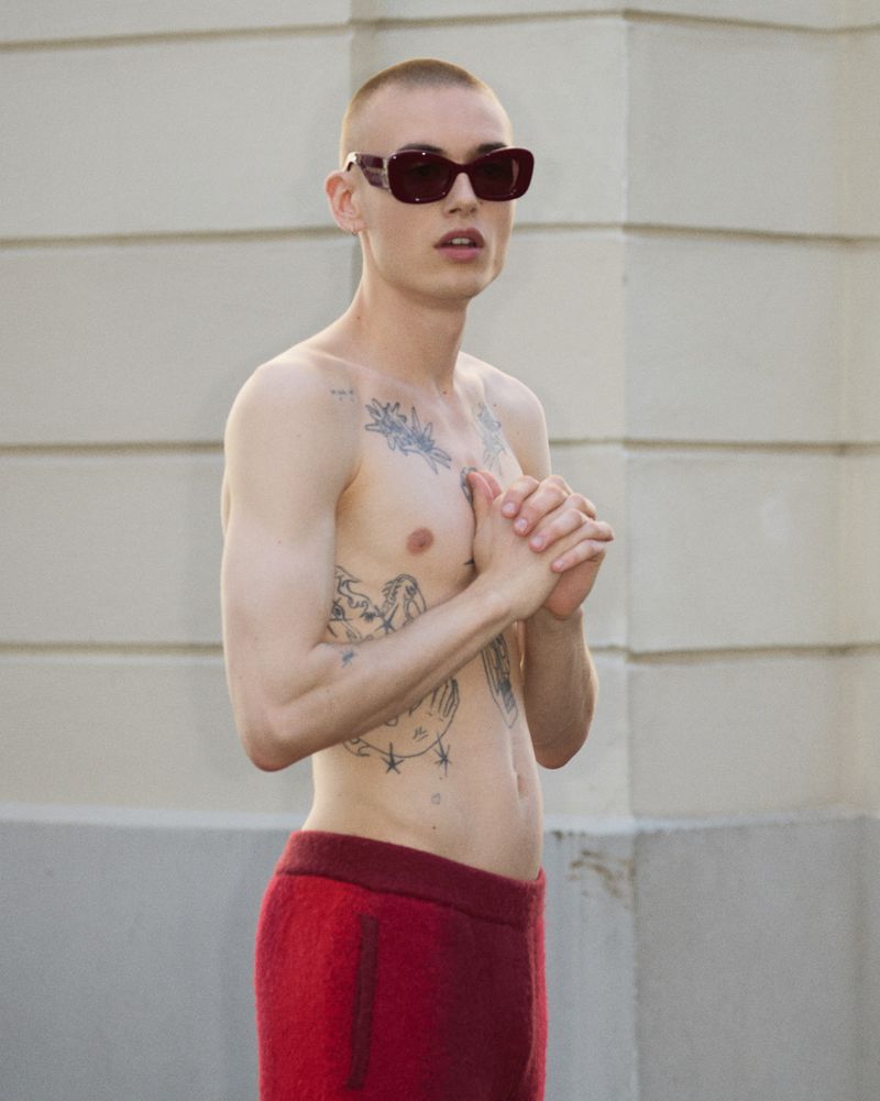 Adult male poses shirtless in red pants and sunglasses outdoors against a light-colored wall, displaying tattoos and a short buzz cut.