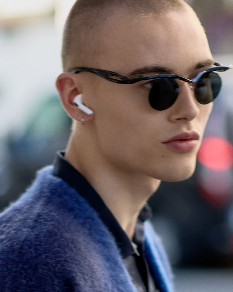 Close-up of a young adult man with sunglasses and wireless earbuds, wearing a textured blue sweater, in an outdoor urban setting.