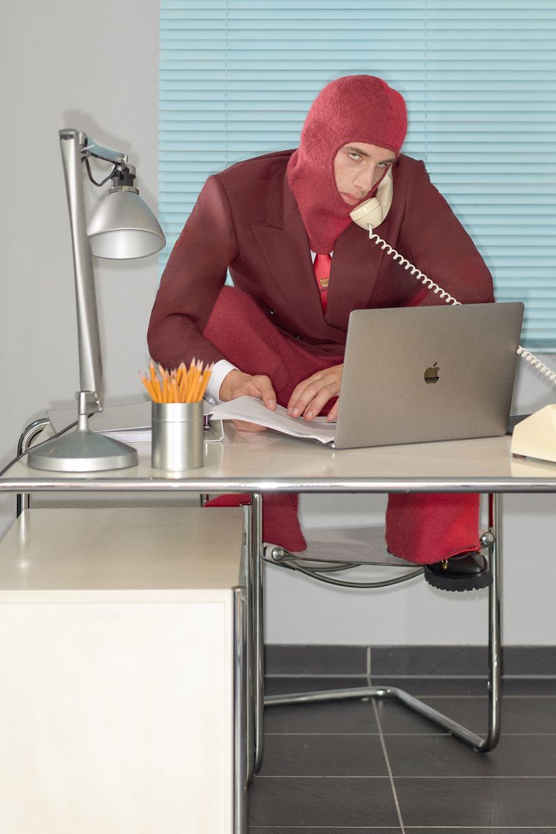 Person in balaclava and suit sits on office desk using laptop and telephone in quirky business fashion editorial, indoor setting.