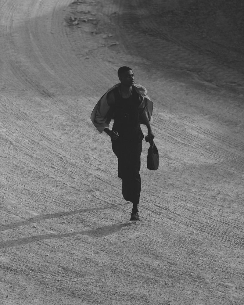 A black and white shot of an adult female walking on a dirt path, carrying a handbag with a light jacket draped over her shoulders.