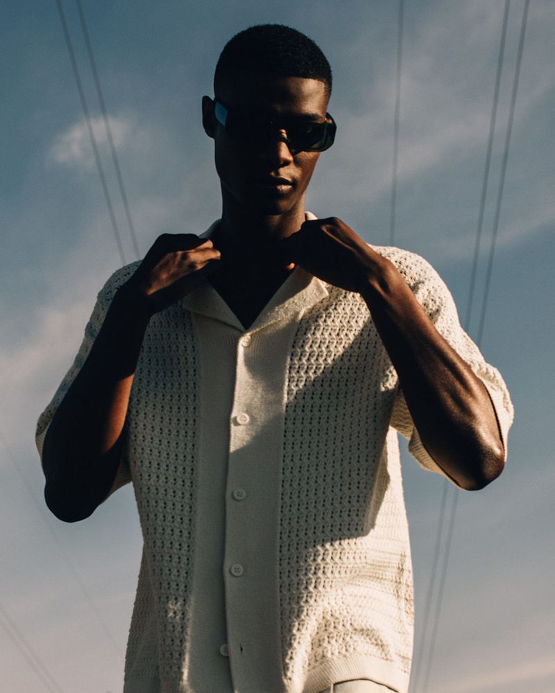 Portrait of an African-American man wearing sunglasses and a button-down knit shirt, posing outdoors with a blue sky and power lines.