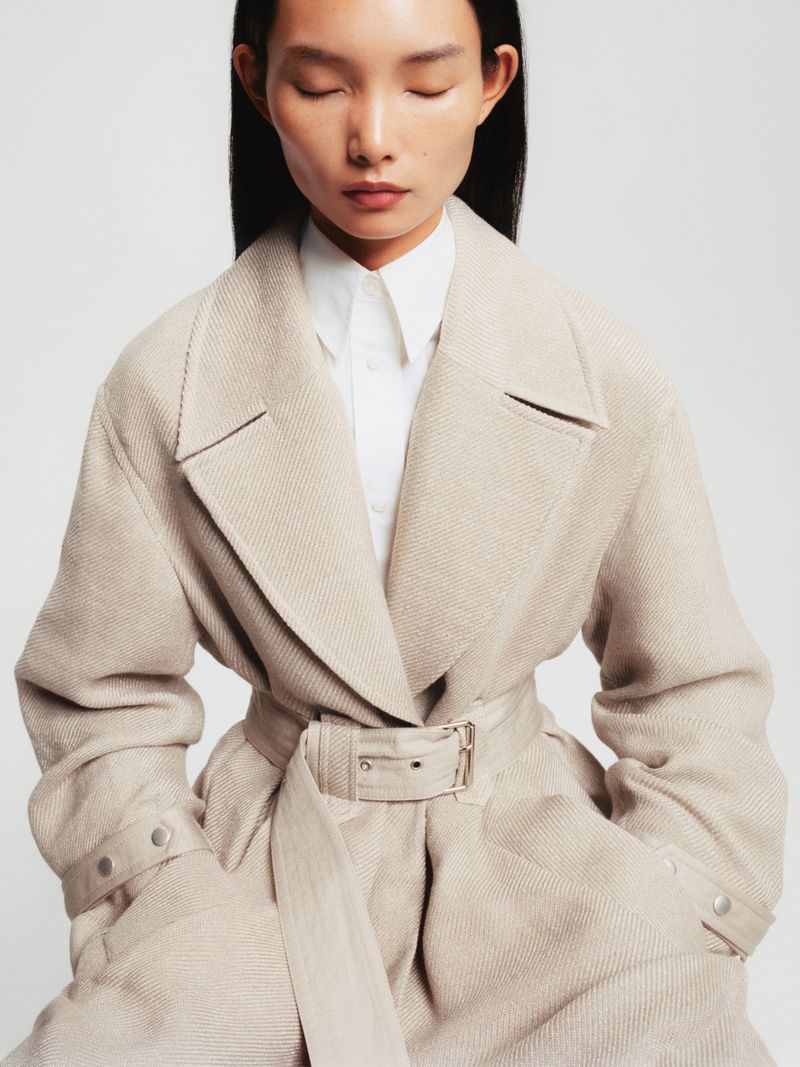 Portrait of an East Asian woman in a beige coat with a belt. The person has their eyes closed and looks peaceful against a white background.