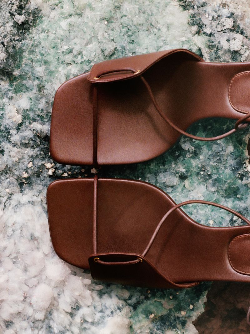 Elevated view of a pair of brown leather sandals with toe loops and delicate ankle straps on a green and white marble surface.