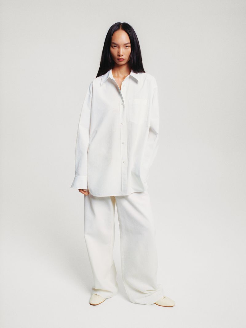 An Asian woman poses in a white button-down shirt and wide-leg pants in a studio setting, creating a clean and minimalist fashion image.