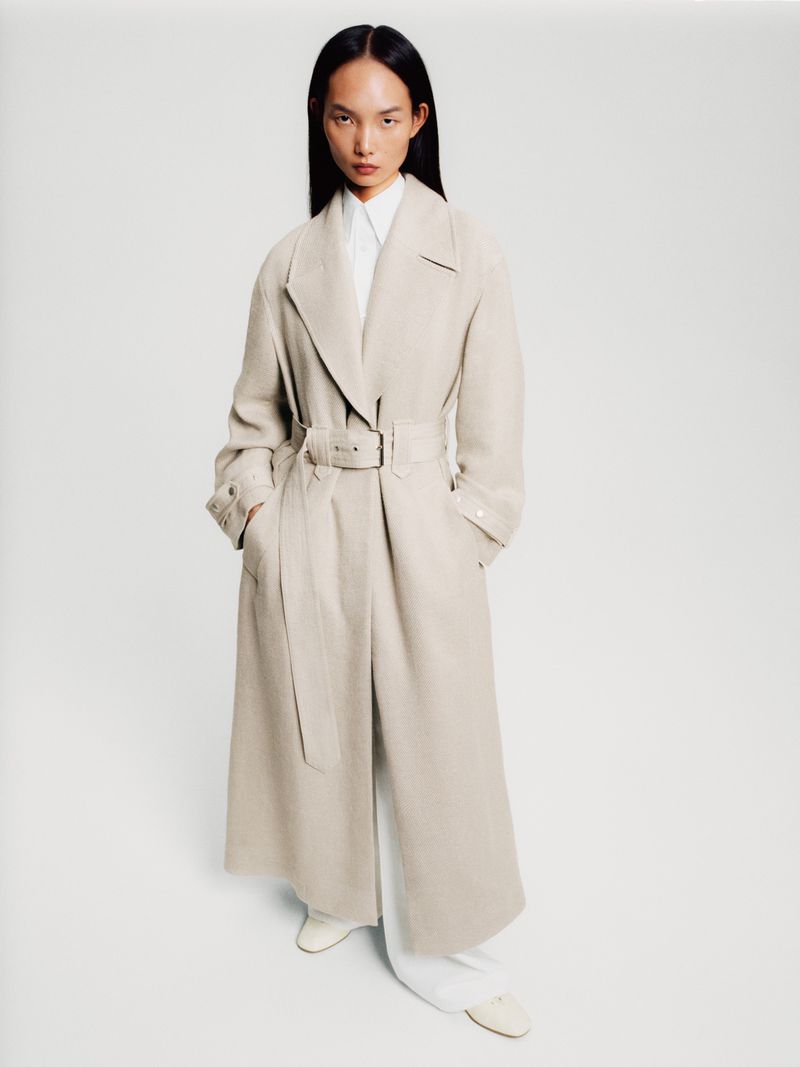 Full-length shot of an adult female model posing in a belted trench coat, long-sleeved white shirt, and white pants in a bright photo studio.