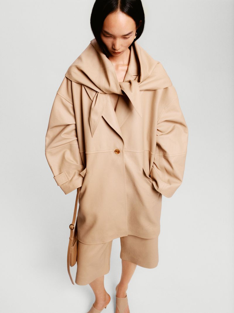 Asian woman poses in a beige oversized coat and matching shorts with a purse on a white background in a fashion shot.