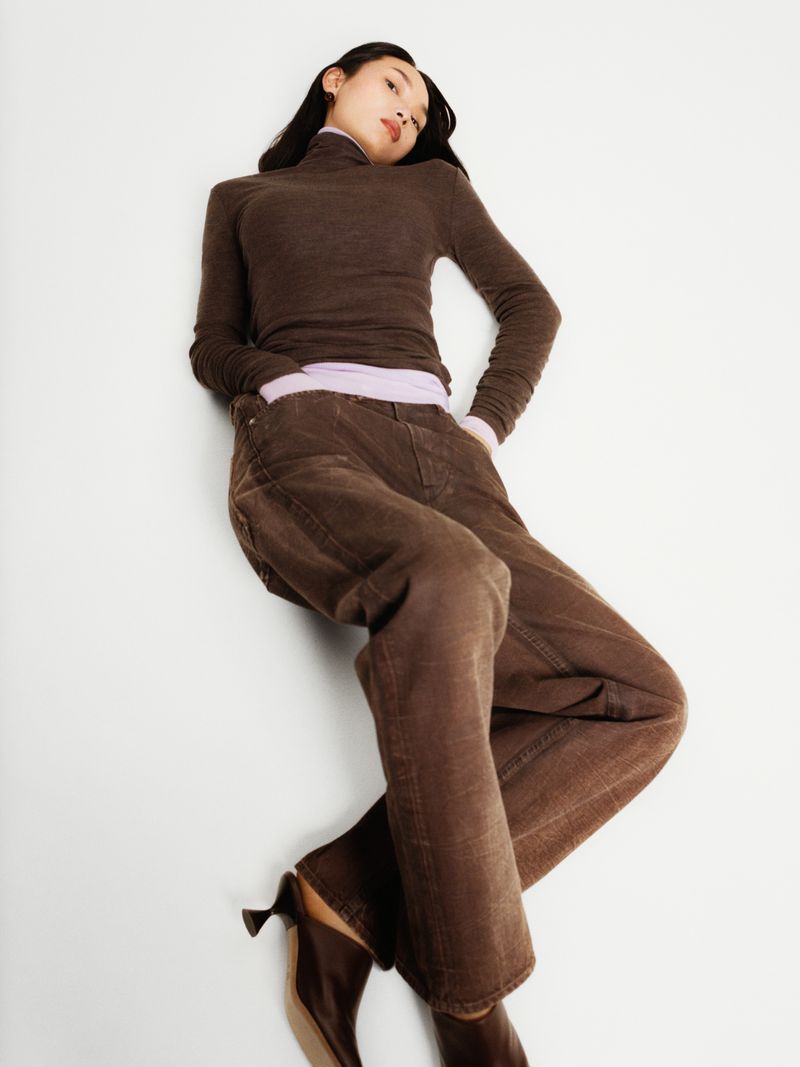 Woman poses in a brown long-sleeved top and brown pants against a white backdrop, showcasing a fashion-forward casual aesthetic.