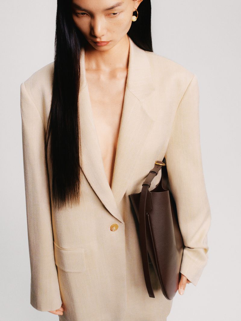 A woman in an oversized beige blazer poses with a dark brown leather purse against a white background in a studio-lit fashion shot.