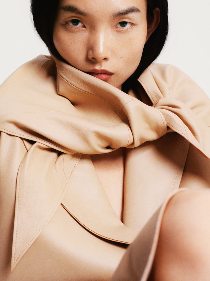 Close-up fashion portrait of an Asian woman with dark hair, wearing a beige trench coat, against a plain background in studio lighting.