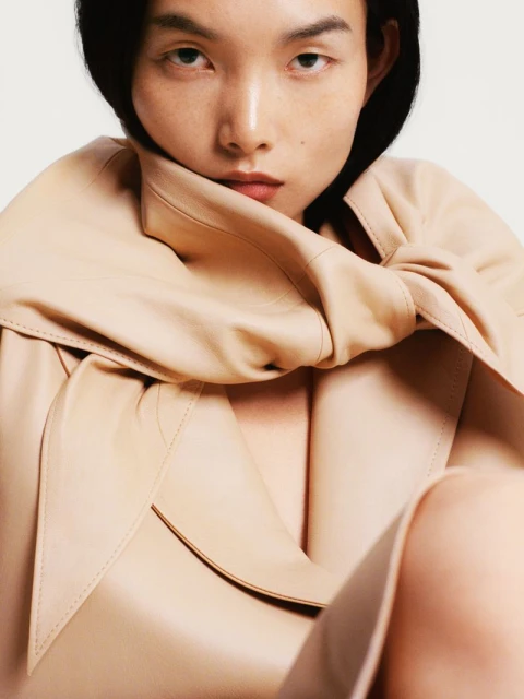 Close-up fashion portrait of an Asian woman with dark hair, wearing a beige trench coat, against a plain background in studio lighting.