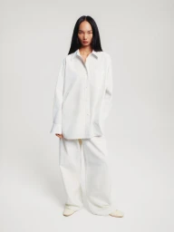 Model Ling Chen wearing an oversized white button-down shirt and wide-leg white trousers against a white seamless studio backdrop.