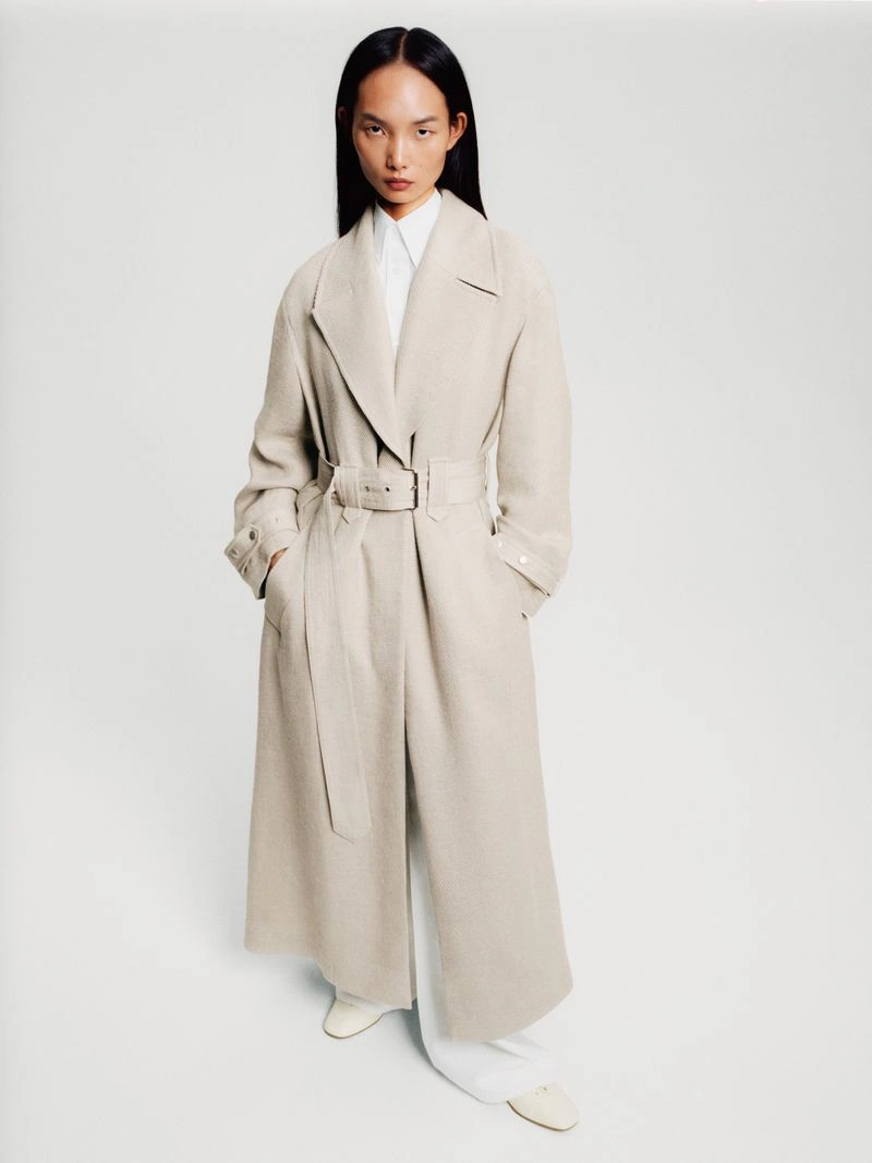 Model Ling Chen wearing a long belted beige trench coat over a white shirt against a plain light studio background.