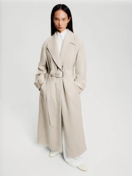 Model Ling Chen wearing a long belted beige trench coat over a white shirt against a plain light studio background.
