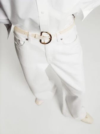 High-angle view of white wide-leg denim trousers, white button-up shirt, and a cream belt with a gold buckle on seamless white backdrop.