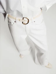 High-angle view of white wide-leg denim trousers, white button-up shirt, and a cream belt with a gold buckle on seamless white backdrop.