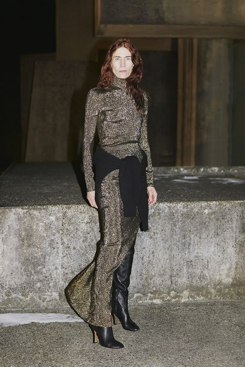 Adult female with red hair in a long gold dress and black boots standing in an outdoor environment with concrete walls and ground.
