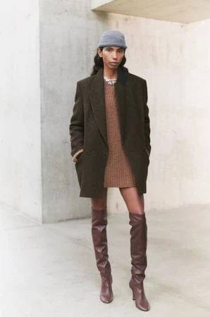 Fashion shot of a woman wearing a brown dress, an oversized brown blazer, and thigh-high brown boots against a neutral background.