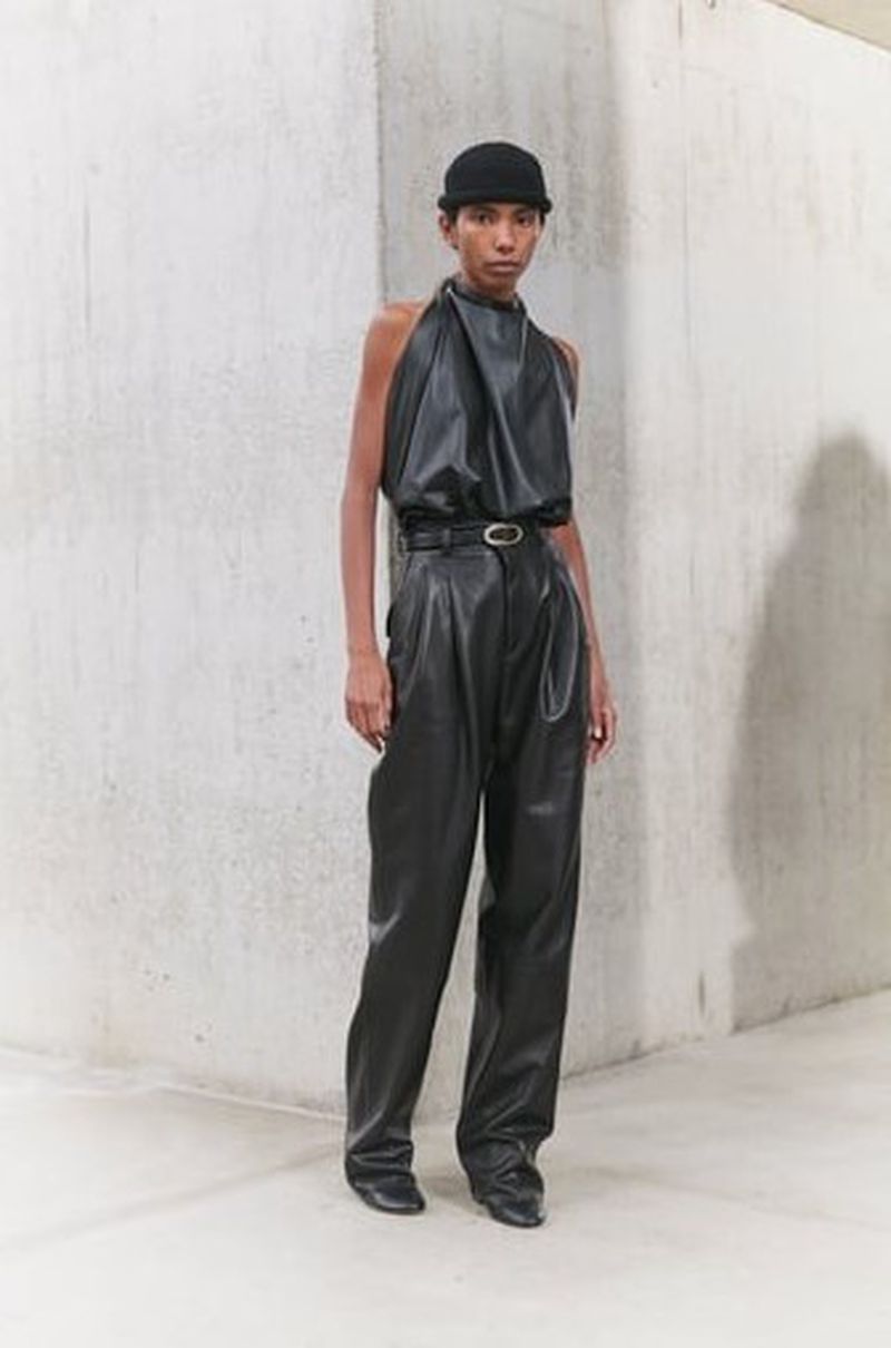A woman in a black leather halter top and high-waisted pants poses indoors against a concrete wall, modeling a chic, modern, edgy fashion look.