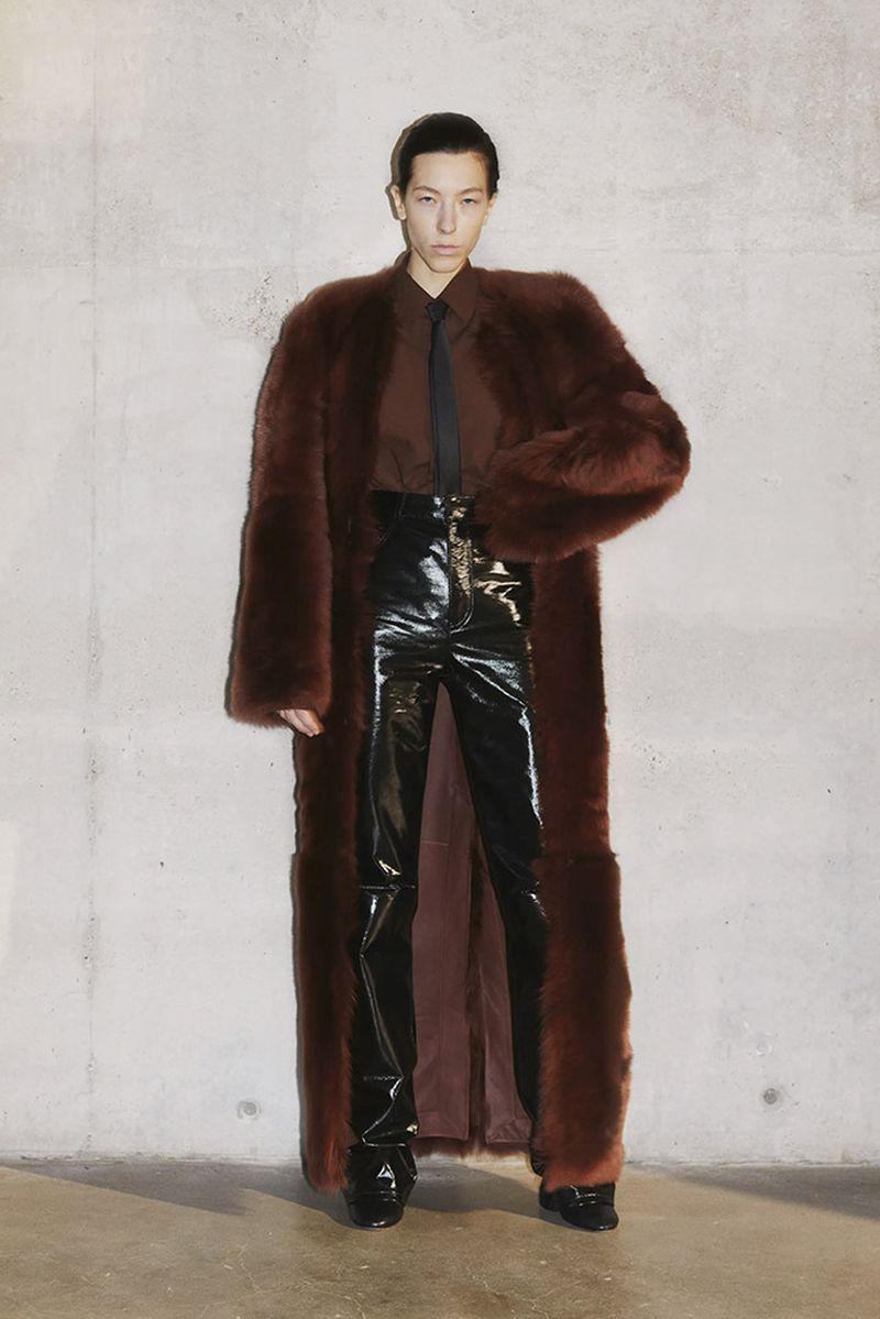 Woman in long brown fur coat, brown shirt, black tie, and high-waisted patent leather pants, posing against a concrete wall.