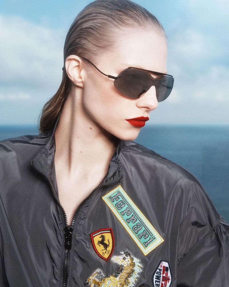 Adult woman with wet hair, red lipstick, and sunglasses wearing a Ferrari jacket in front of a blue sky and ocean background.