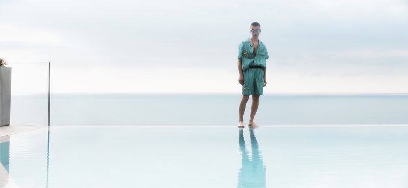A man stands on the edge of an infinity pool with the ocean and sky in the background wearing a matching teal patterned shirt and shorts.