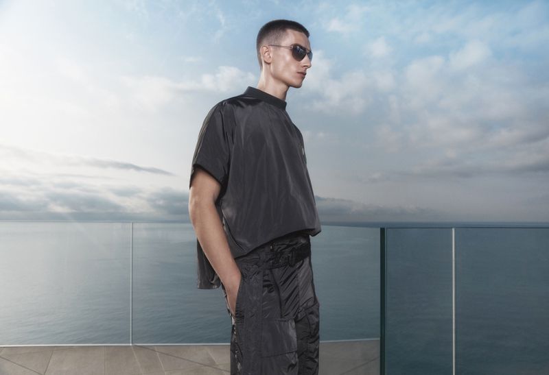 A fair-skinned adult male model stands on a balcony overlooking the ocean, wearing sunglasses and dark, modern, high-fashion, loose-fitting clothing.