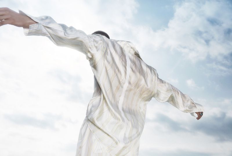 Model with arms outstretched viewed from behind against a cloudy sky wearing a pale patterned silk shirt.