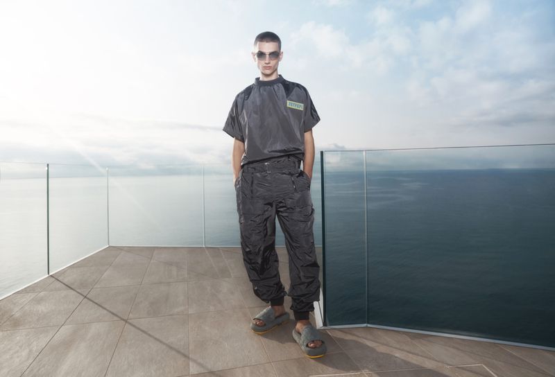 A stylish adult male in a grey outfit poses on a rooftop with a sea view, wearing sunglasses and slides. Fashion editorial shot.