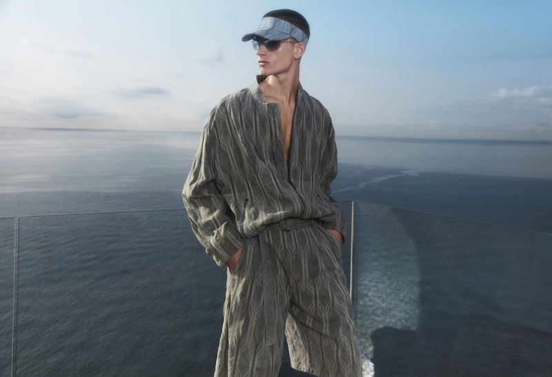 Fashion shot of a man in a textured linen shirt and shorts, visor and sunglasses posing in front of the ocean and blue sky.