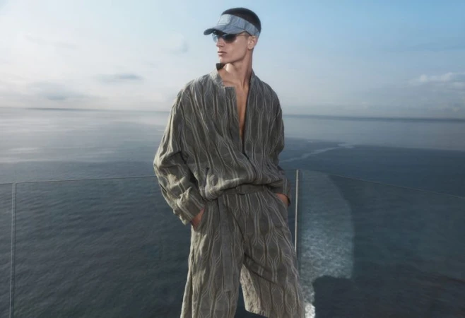 Fashion shot of a man in a textured linen shirt and shorts, visor and sunglasses posing in front of the ocean and blue sky.