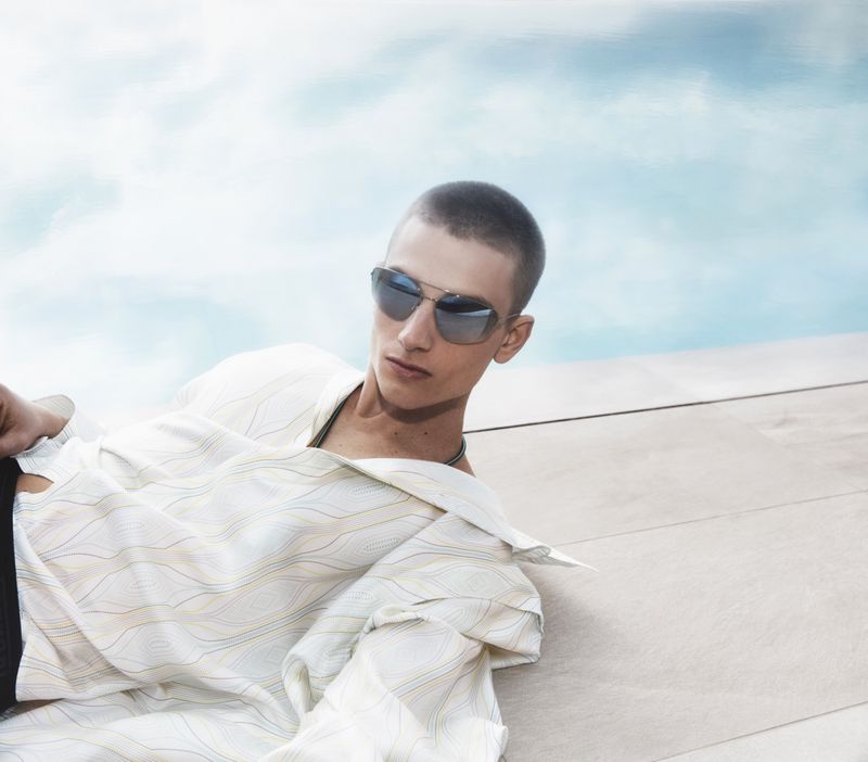 Model with a buzz cut wearing aviator sunglasses and a cream patterned shirt reclining poolside.