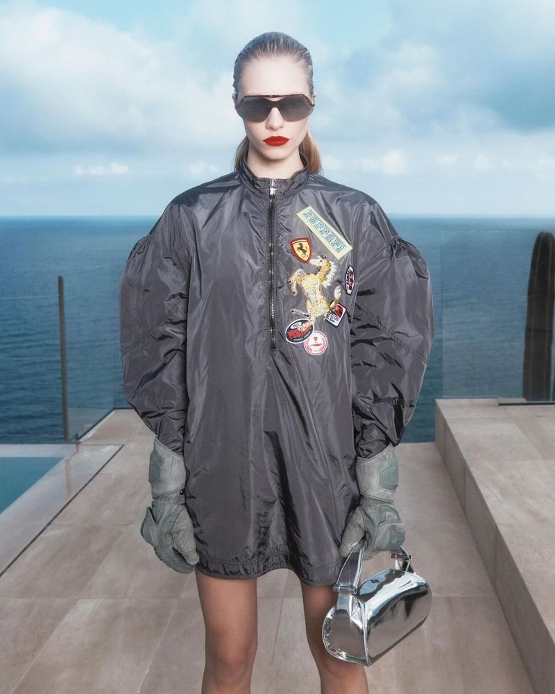 Woman in oversized bomber jacket, gloves, and sunglasses stands outdoors with a metallic handbag, ocean, and cloudy sky backdrop.