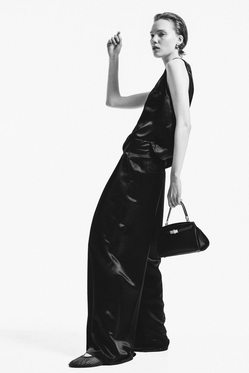 A woman in a black satin top and pants holds a black purse in a studio setting, creating a high-contrast black and white fashion portrait.