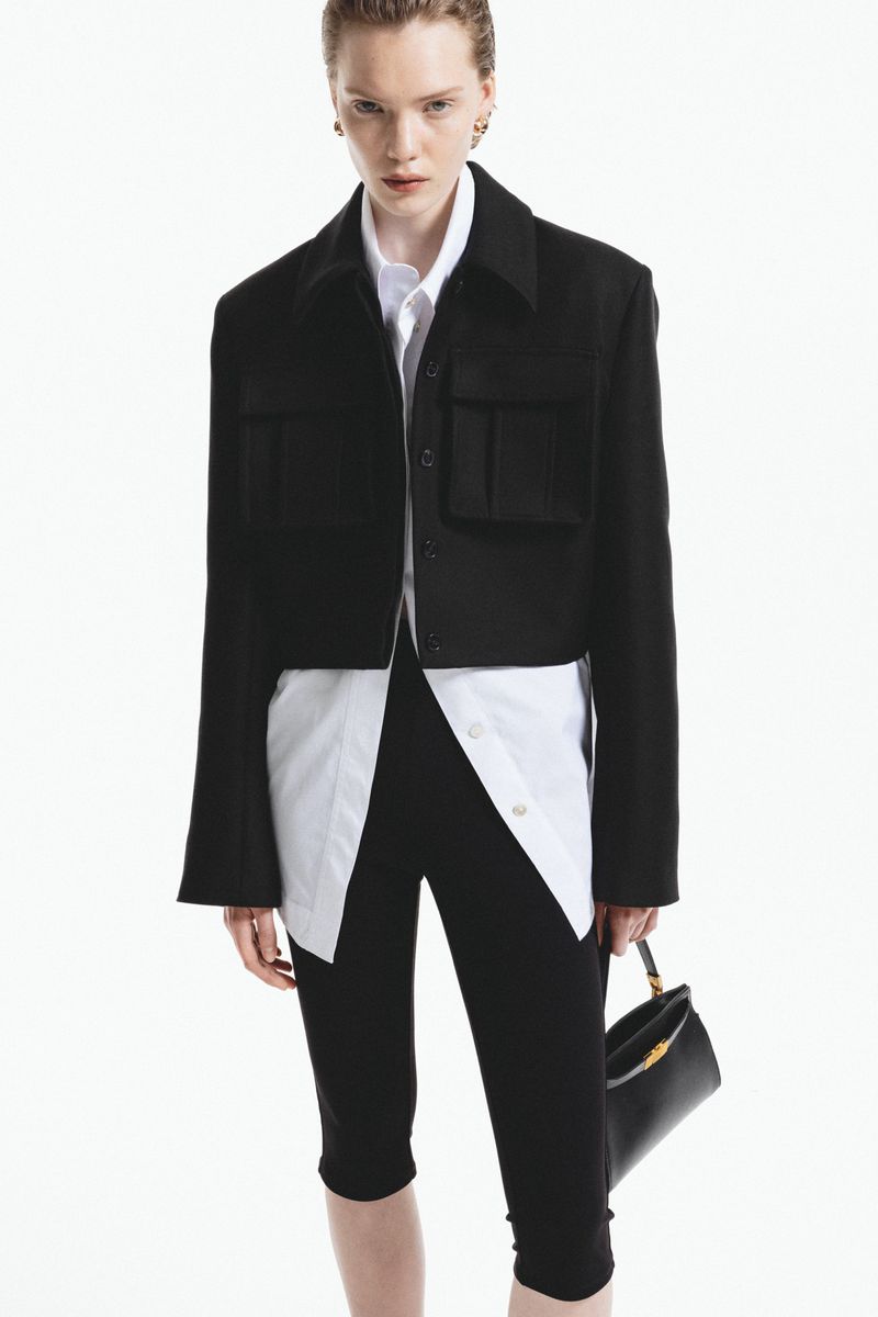 Fashionable woman in black jacket and leggings with white shirt, holding a stylish handbag against a clean white background.