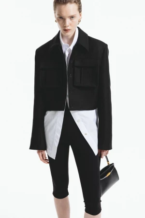 Fashionable woman in black jacket and leggings with white shirt, holding a stylish handbag against a clean white background.