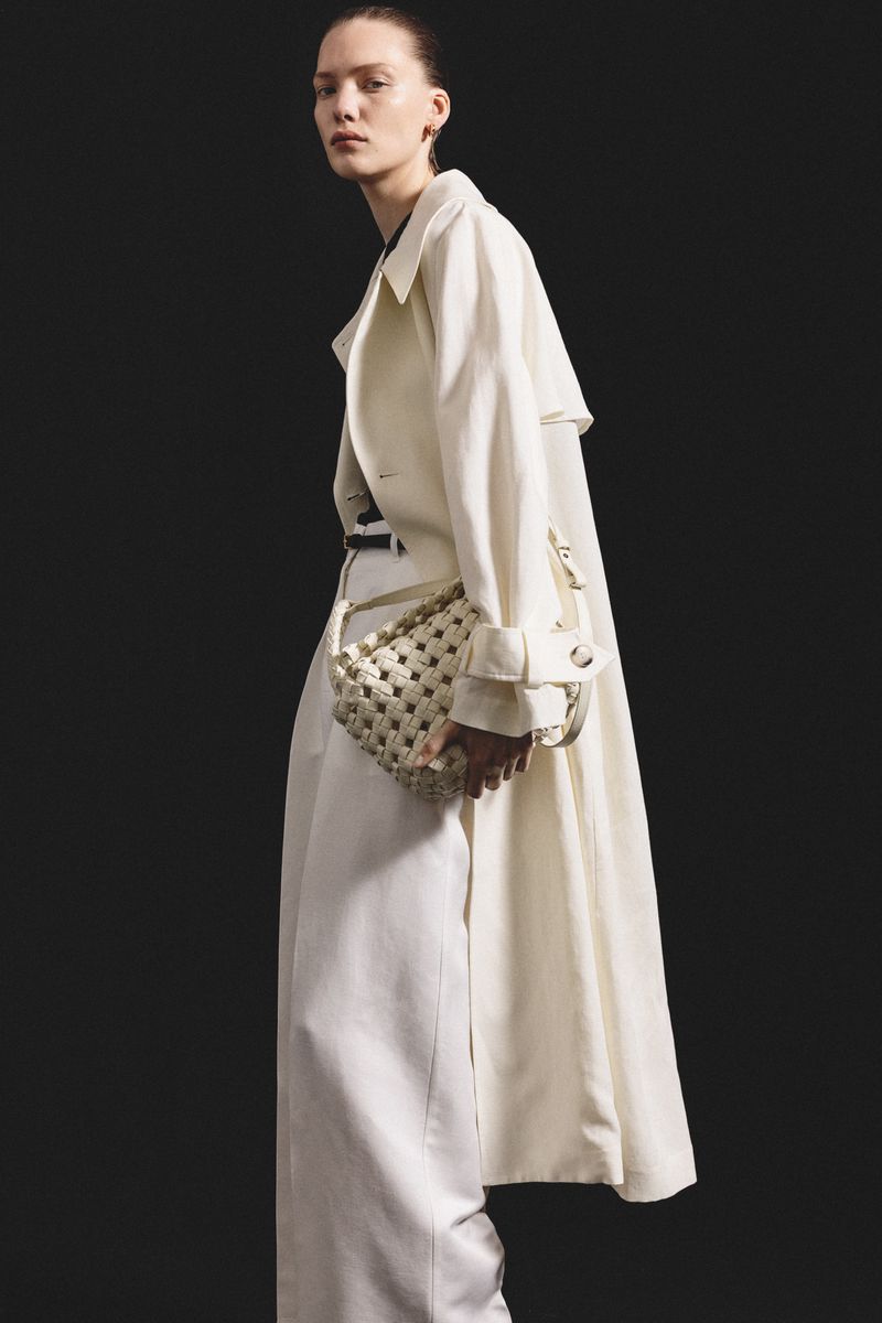 Fashion model in a studio wearing a white trench coat and wide-leg trousers, holding a woven handbag, against a dark background.