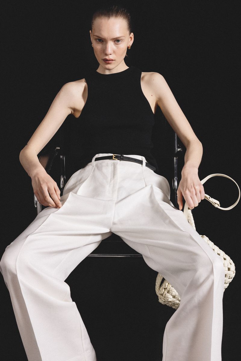 Studio shot of a young adult woman in a black tank top and white wide-leg pants, seated in a modern chair with a woven bag.