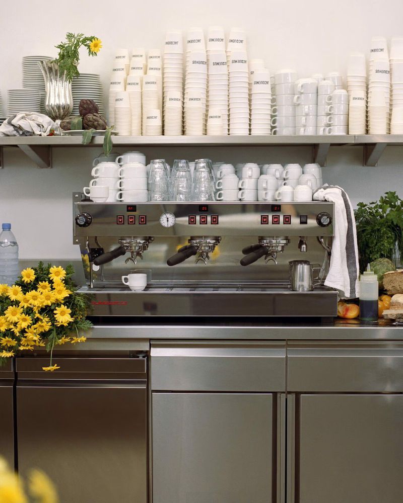 Espresso machine and stacks of cups on stainless steel shelves in a café, shot in eye-level medium indoor shot with natural lighting.
