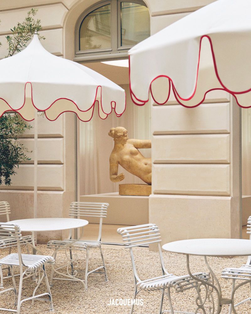 Outdoor courtyard with white metal chairs and tables under white umbrellas with a statue and building in the background. 