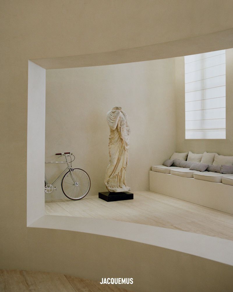 Interior view with a white bicycle, a classical sculpture on a black base, window light, and a low bench with pillows.
